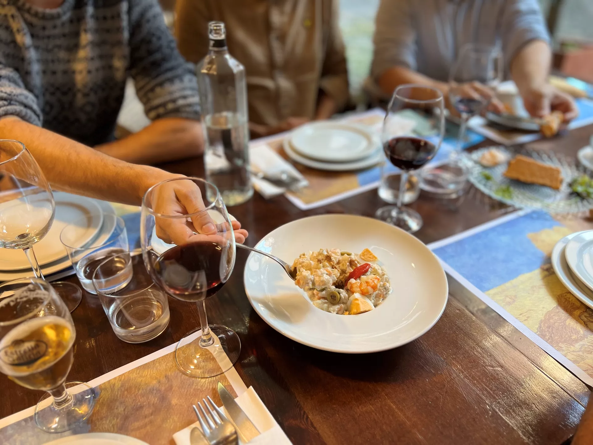 Mesa de restaurante con varios comensales, comida y bebida servida, mientras uno de los comensales se sirve ensaladilla rusa